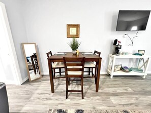 Een eetgedeelte met een houten tafel, vier stoelen, een kleine plant, een lijst met een foto en een aan de muur bevestigde televisie.
