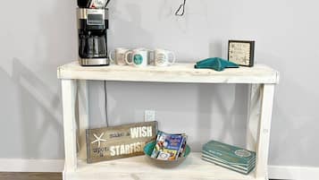 A white wooden table with a coffee maker, three mugs, a starfish decoration, a sign, a bowl of magazines, and a stack of books.