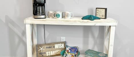 A white wooden table with a coffee maker, three mugs, a starfish decoration, a sign, a bowl of magazines, and a stack of books.