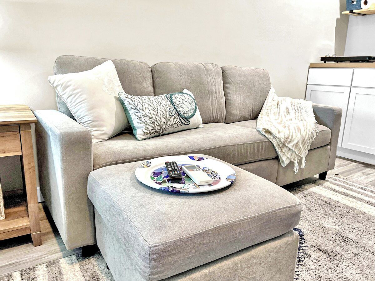 A living room with a grey sofa, a small ottoman, and a coffee table with remote controls and a phone.