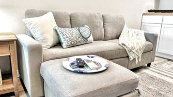 A living room with a grey sofa, a small ottoman, and a coffee table with remote controls and a phone.