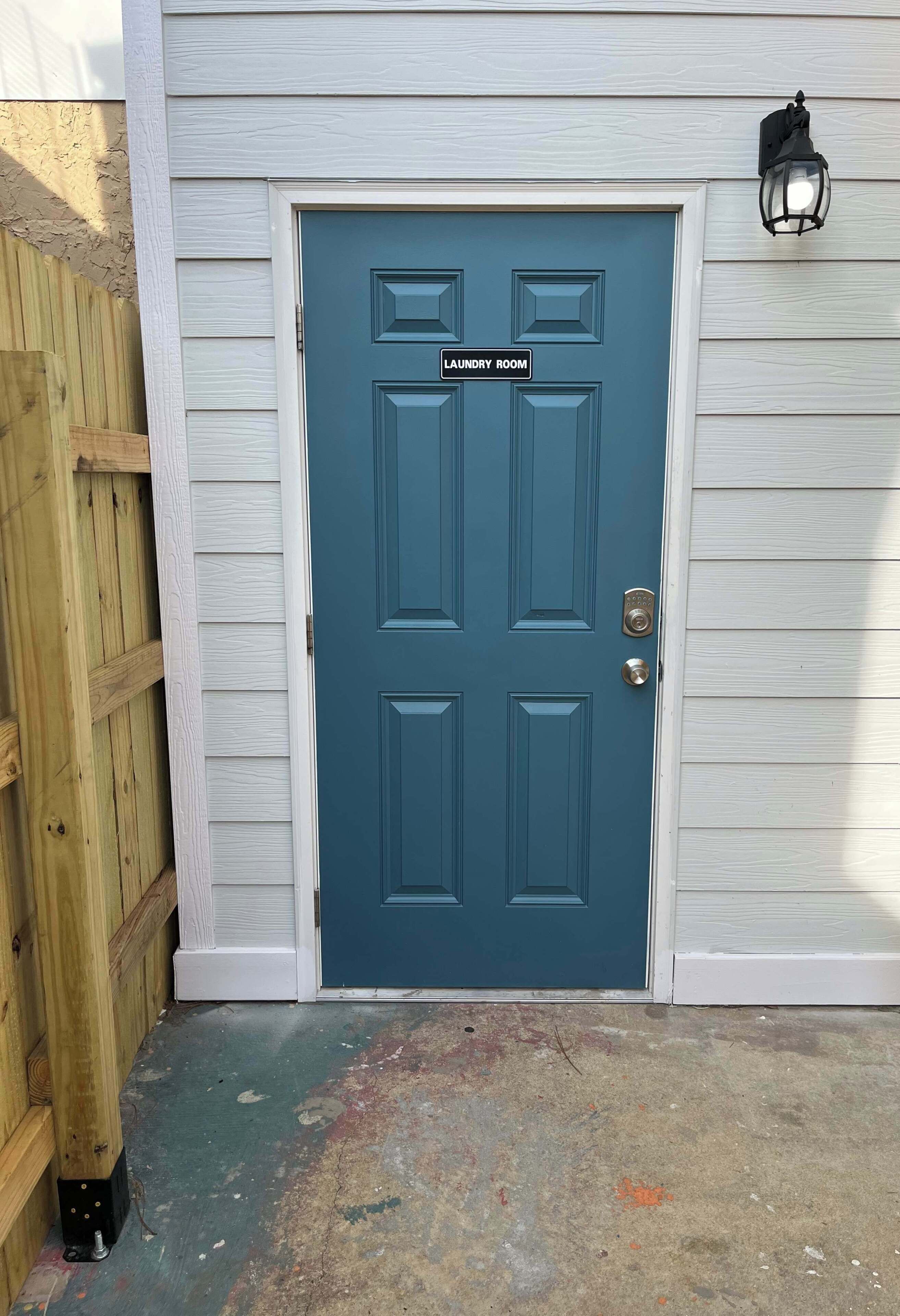 A dark blue door with a sign indicating a laundry room, a wooden fence to the left, and a light-colored exterior wall.