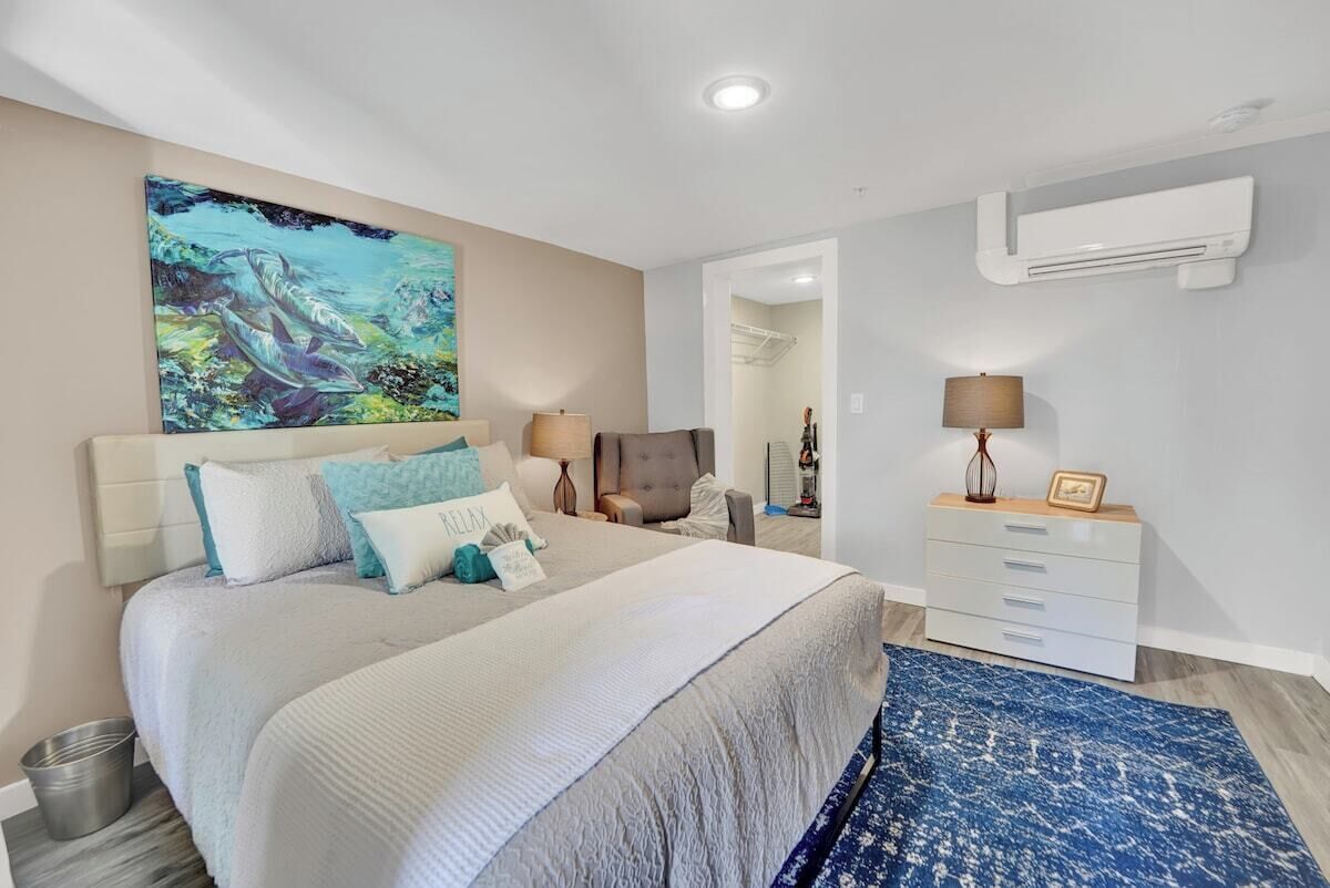 A bedroom with a bed, bedside tables, a nightstand, a painting, and an air conditioning unit.