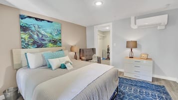 A bedroom with a bed, bedside tables, a nightstand, a painting, and an air conditioning unit.