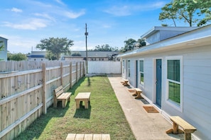 A fenced backyard with a grassy area, wooden benches, and a building with windows. - Seaside Escape 7 - Relaxing Coastal Escape (Fort Walton Beach)