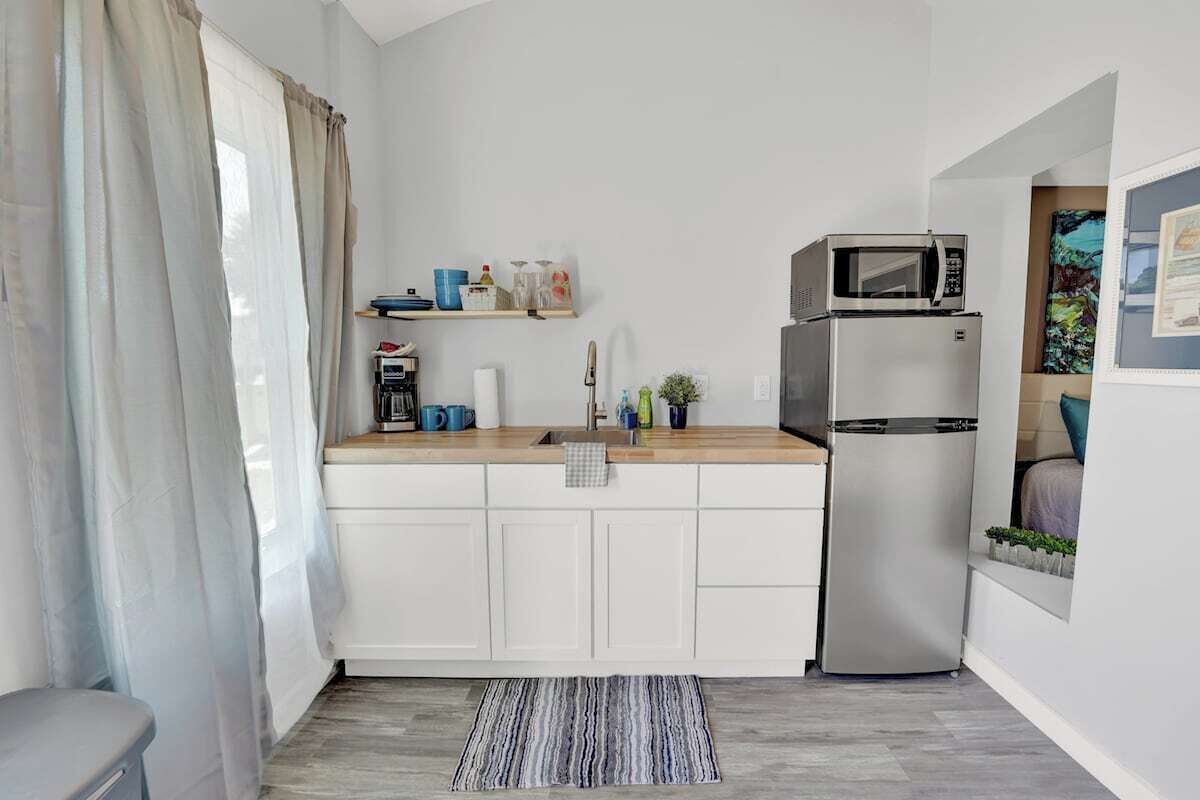 A compact kitchen with white cabinets, a microwave, a refrigerator, and a small shelf with kitchen items.