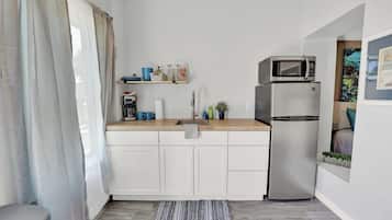 A compact kitchen with white cabinets, a microwave, a refrigerator, and a small shelf with kitchen items.