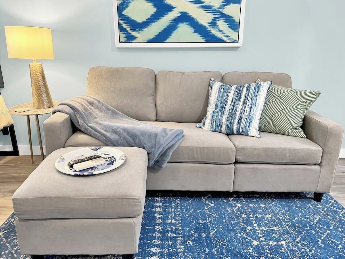 Una sala de estar con un sofá color crema, un tapete estampado azul y una mesita de noche con una lámpara y controles remotos.