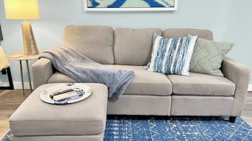 A living room with a beige sofa, a blue patterned rug, and a side table with a lamp and remote controls.