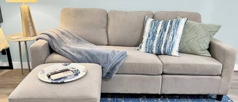 Una sala de estar con un sofá color crema, un tapete estampado azul y una mesita de noche con una lámpara y controles remotos.