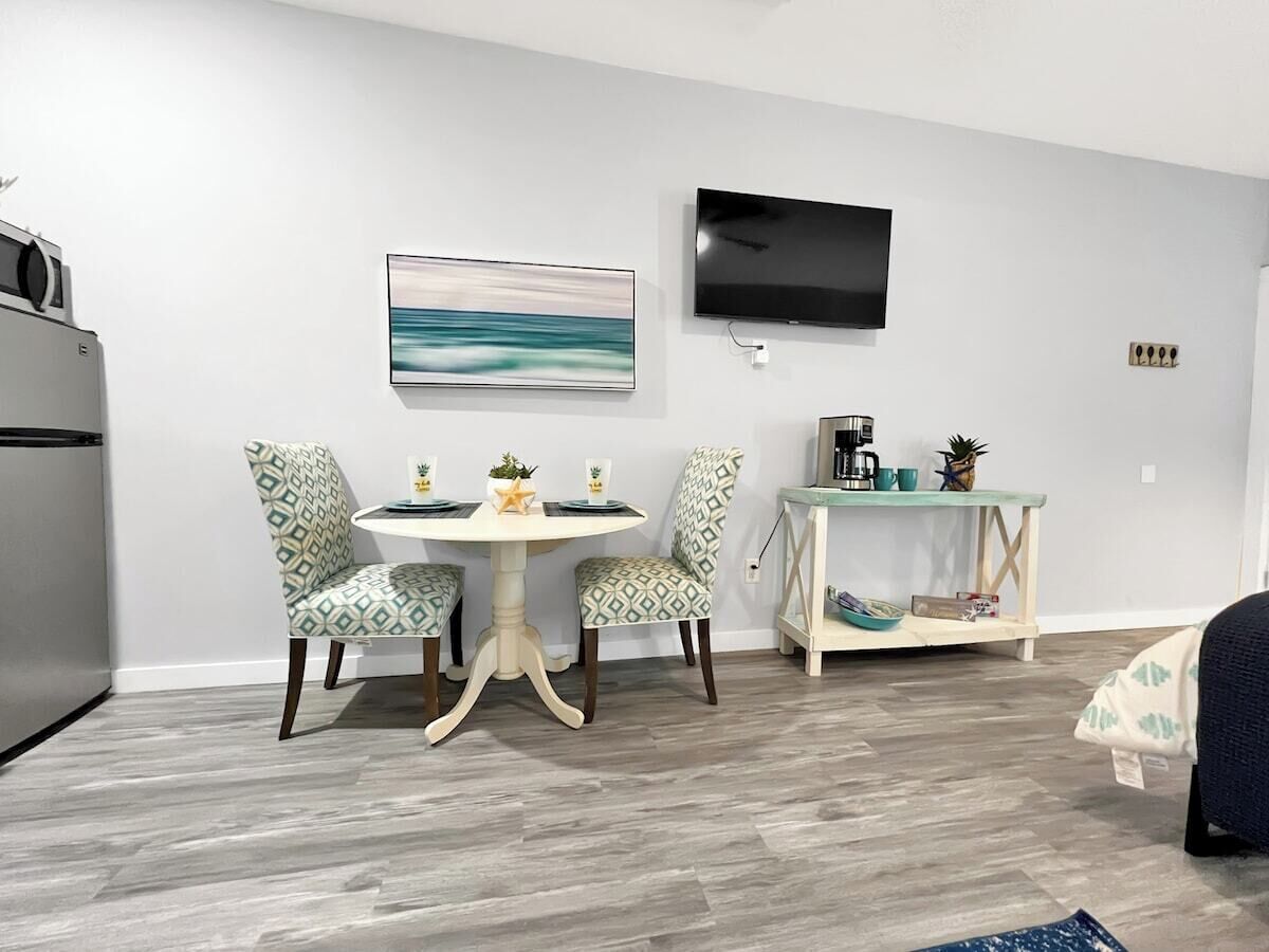 A modern kitchen with a built-in oven, a dining area with patterned chairs, a round table, and a flat-screen TV mounted on the wall.