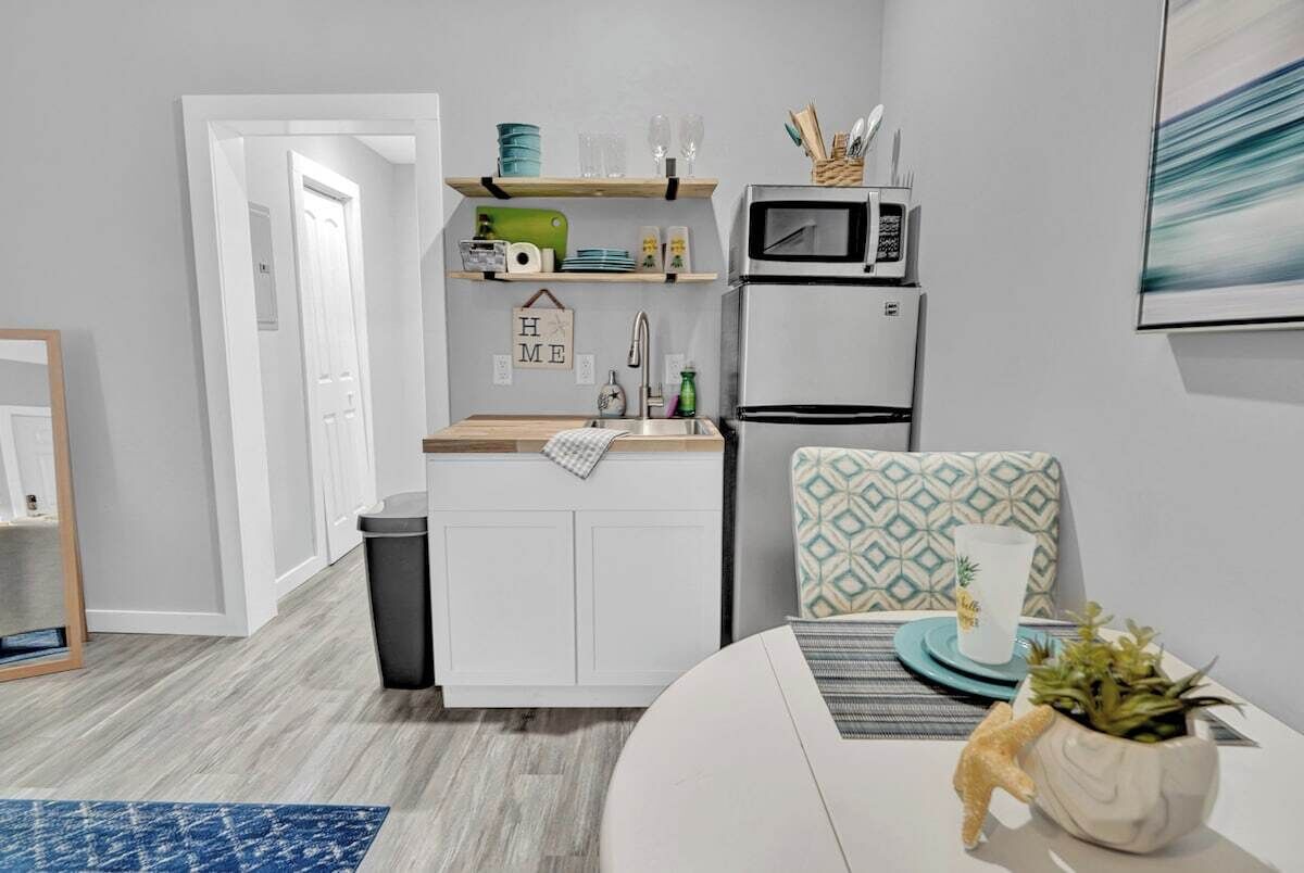 A compact kitchen with a microwave, a sink, and a small countertop. There is a trash bin and a chair with a patterned cushion.