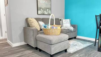 Un salon avec un canapé gris, un pouf assorti, un panier en osier rempli de livres et une œuvre d’art encadrée accrochée au mur.