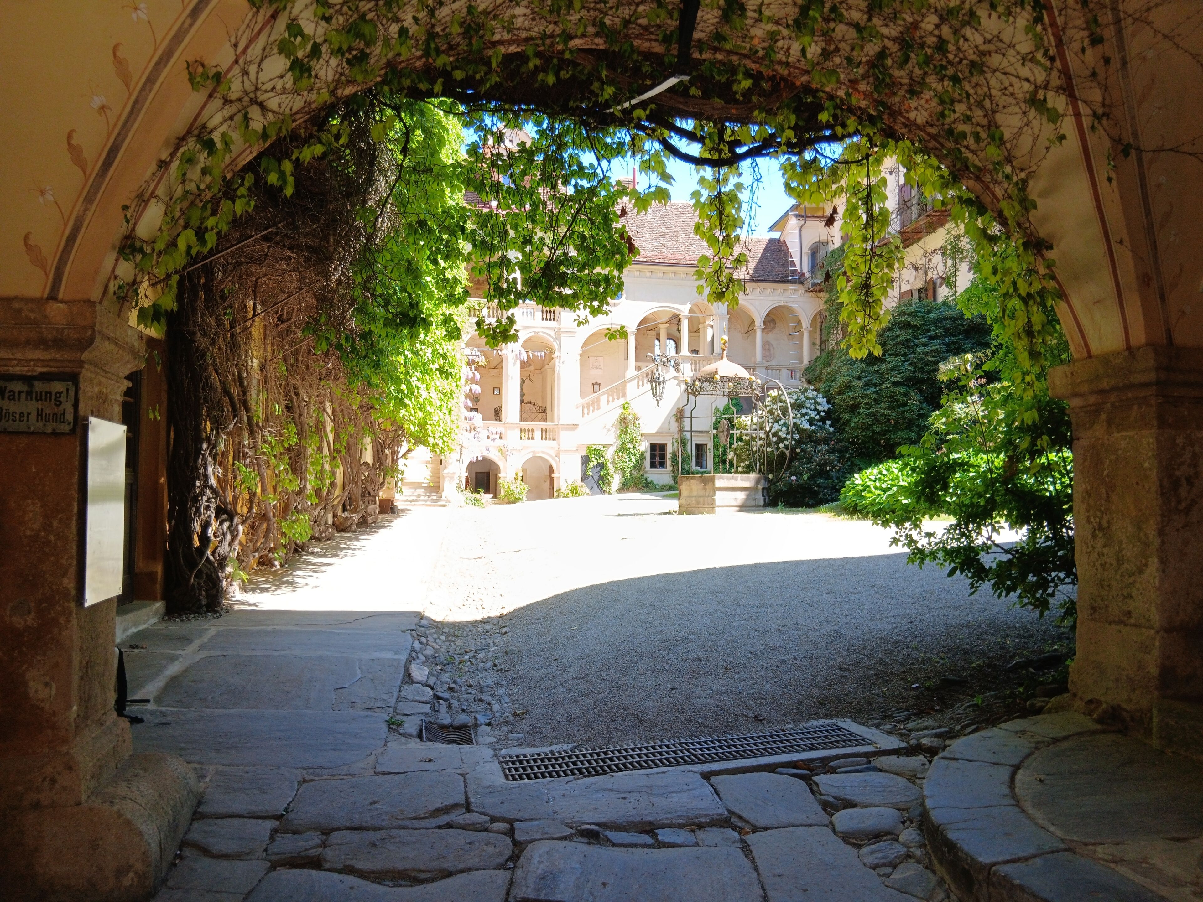 An arched stone pathway leading to a courtyard with a fountain and a building in the background.
