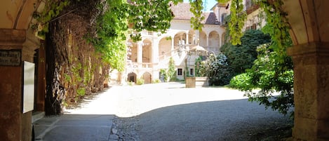 An arched stone pathway leading to a courtyard with a fountain and a building in the background.