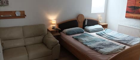 Quarto com cama, sofá, mesa de cabeceira com luminária, relógio e janela.