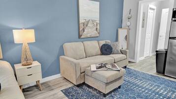 A living room with a sofa, ottoman, and a lamp on a nightstand.