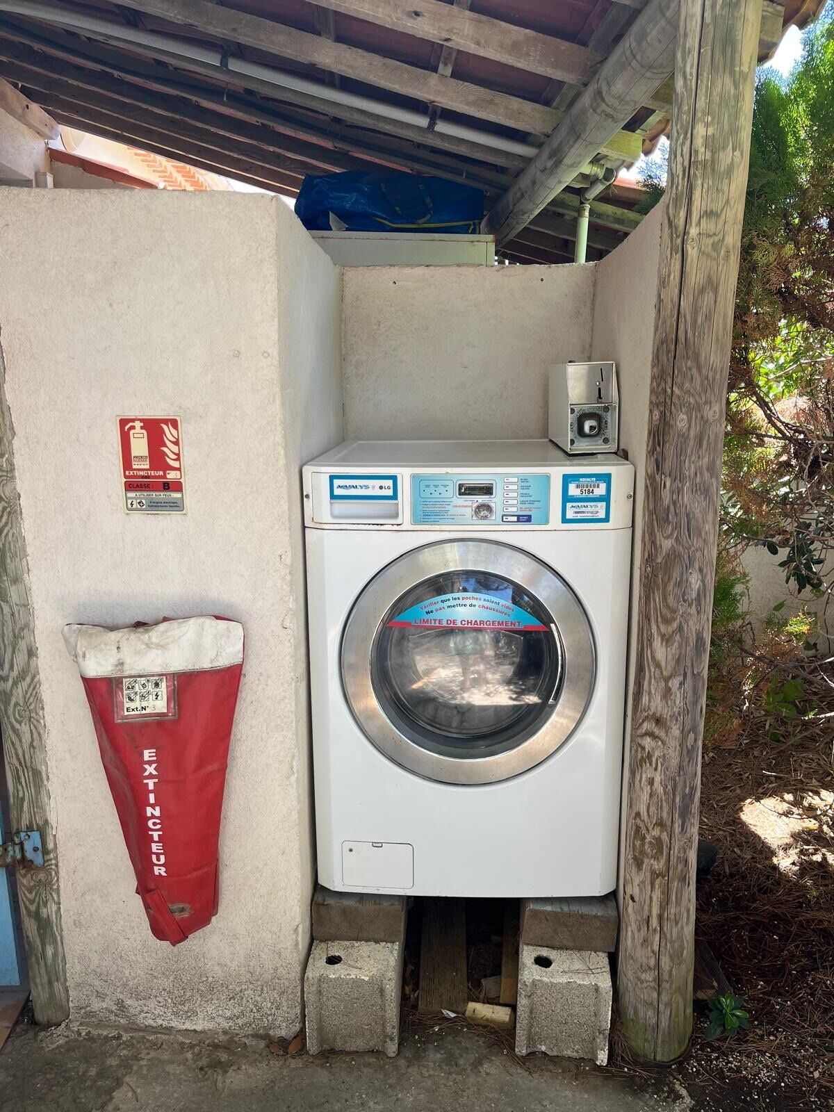 A compact laundry facility with a front-loading washing machine, a red bag labeled 'EXTINTEUR,' and a small control panel.