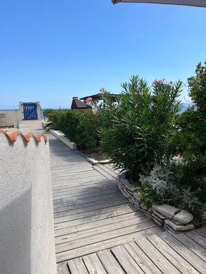 Una terraza de madera que conduce a un edificio con un portón azul, rodeado de follaje y un cielo despejado.