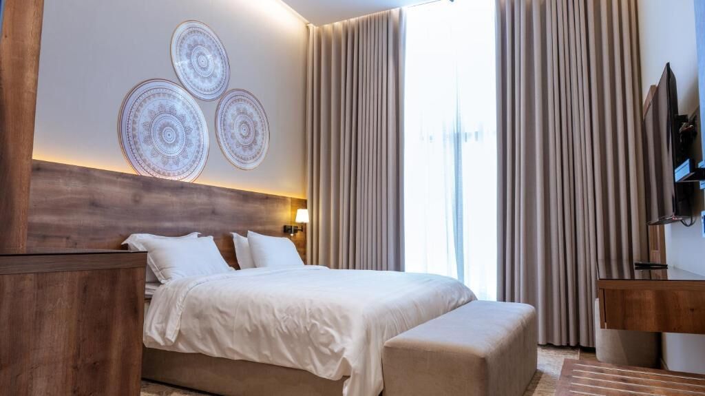 A hotel room with a bed, a wooden headboard, a bedside table, a TV, and curtains.