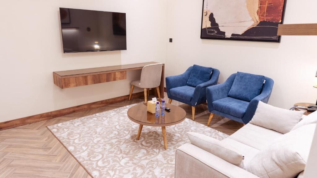 En moderne stue med et fladskærms-tv på væggen, et træ-konsolbord, to blå lænestole, en hvid sofa og en gulvtæppe med et mønster.