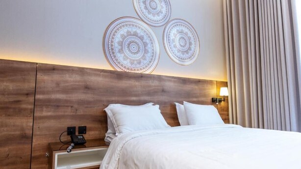 A hotel room with a wooden headboard, a bed with white bedding, a bedside table with a phone, and a wall-mounted lamp.