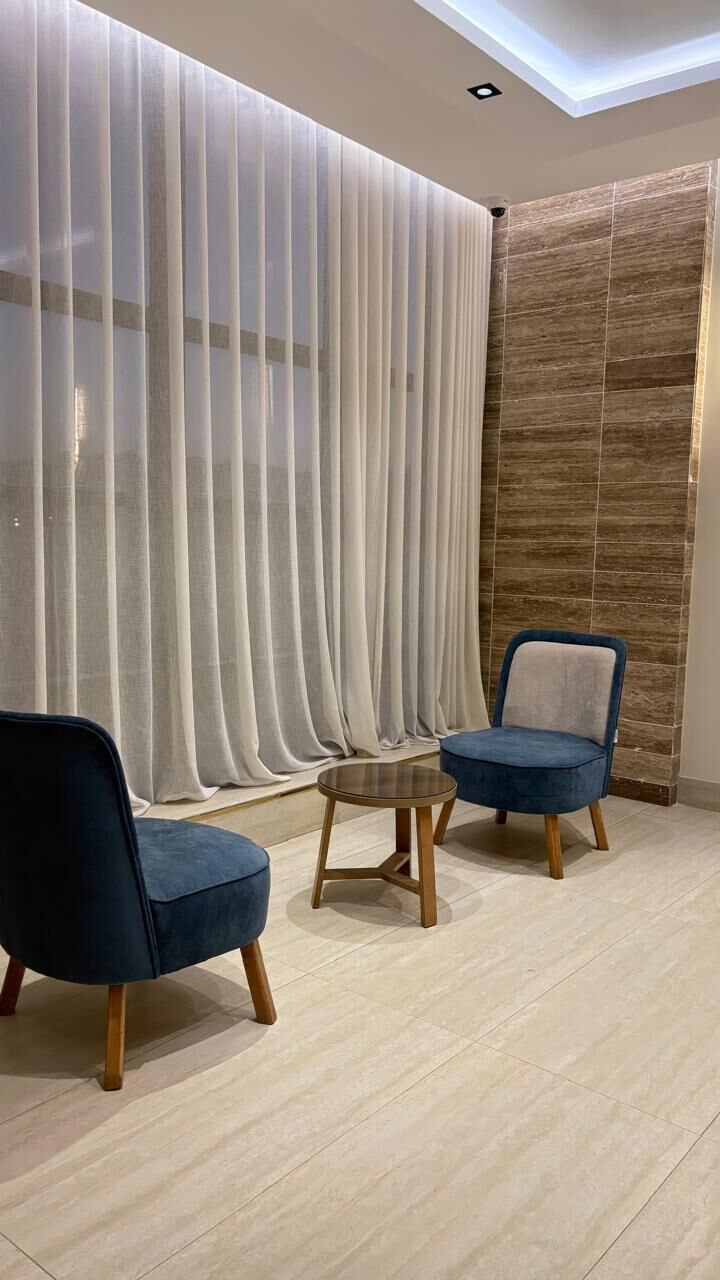 Et værelse med moderne design, med et stort vindue med lette gardiner, et siddeområde med to stole og et lille bord samt en væg med struktur.