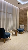 Una habitación con diseño moderno, que incluye un amplio ventanal con cortinas translúcidas, un rincón de descanso con dos sillones y una mesita, y una pared con textura.