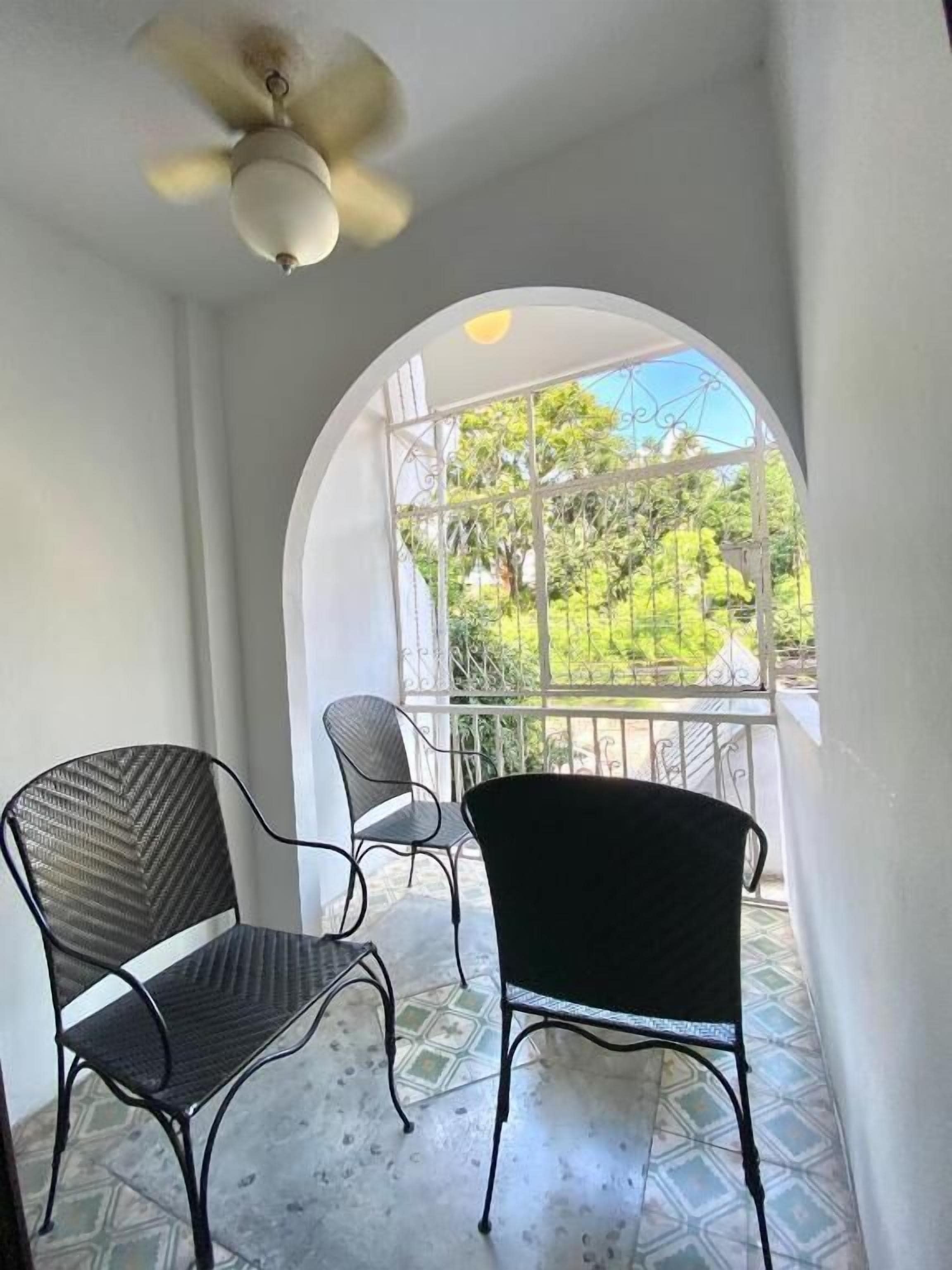 A covered patio with two chairs, a ceiling fan, and a view of greenery through an arched window.