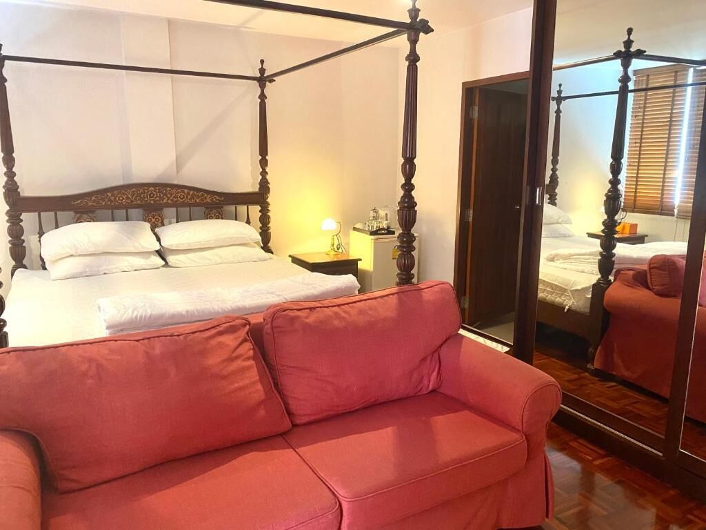 Una cama con dosel, un sofá rojo, una mesita de noche con lámpara y un espejo que refleja la habitación.