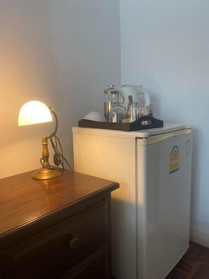 Un refrigerador compacto con cubierta de vidrio, una lámpara sobre una mesita de noche de madera, y una repisa en la pared con utensilios de cocina.