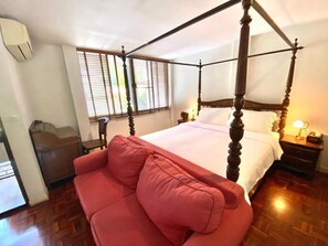 Una cama con dosel y estructura de madera, un sofá rojo, una mesita de noche con una lámpara y una ventana con persianas.
