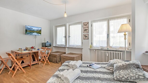 A well-lit room with a bed, a desk, and a chair. There are windows with curtains, a radiator, and a lamp. - Gran Roc 9 (Sestriere)