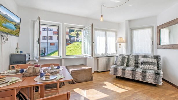 Un salón luminoso con un sofá, una mesa de comedor con sillas y vistas a un edificio a través de amplios ventanales.