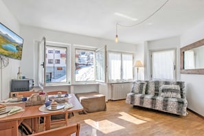 Ein gemütliches Wohnzimmer mit Holzboden, einer Couch, einem Esstisch mit Stühlen und einem Fernseher, der eine verschneite Landschaft zeigt.