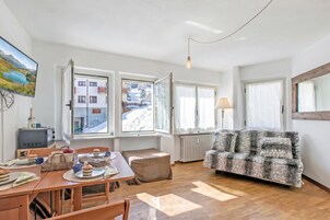 A cozy living room with a wooden floor, a sofa, a dining table with chairs, and a television displaying a snowy landscape.