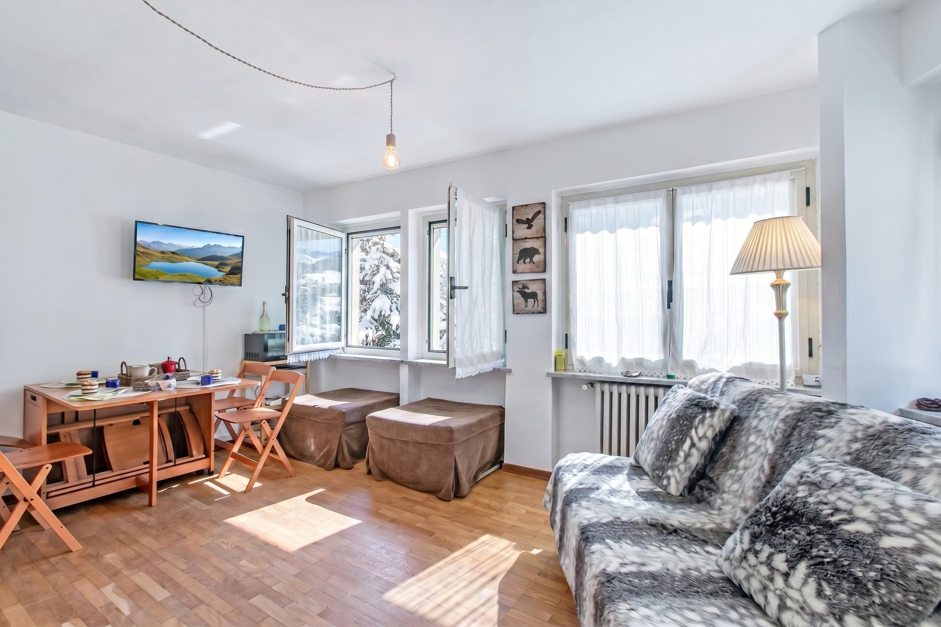 A living room with a wooden floor, a sofa, a TV, a dining table with chairs, and a window with curtains.