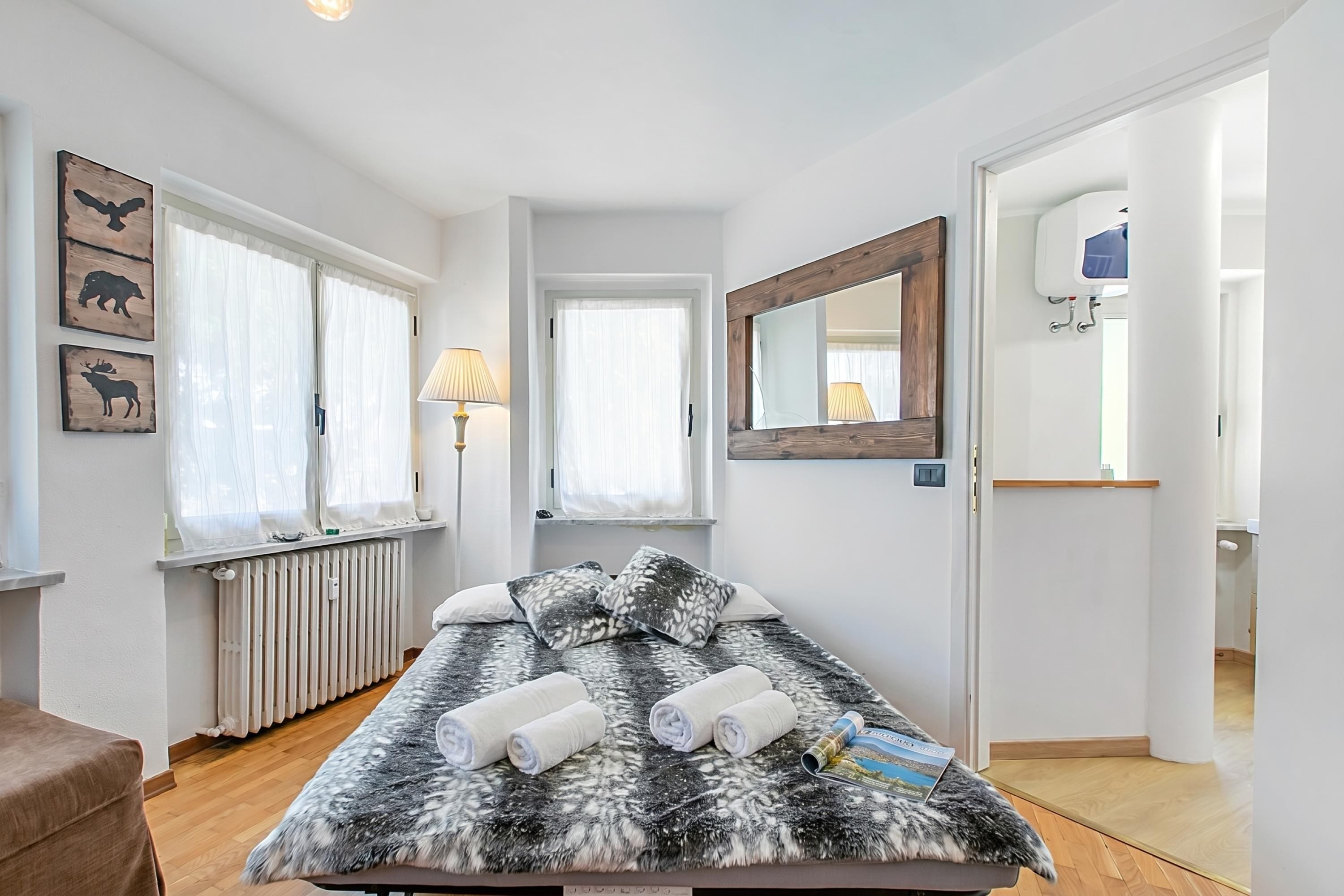 A bedroom with a bed, two bedside lamps, a radiator, a window with curtains, a mirror, and a door leading to a bathroom.