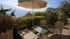 An outdoor seating area with a table and chairs, an umbrella, and a view of the sea and mountains in the background. - Arias Giovanna Visa Lago Villa (Tremosine)