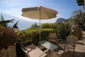 Ein Sitzbereich im Freien mit Tisch und Stühlen, einem Sonnenschirm und Blick auf Meer und Berge im Hintergrund.