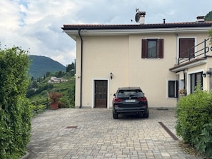 A house with a paved driveway, a parked car, and a mountain in the background. - Arias Giovanna Visa Lago Villa (Tremosine)