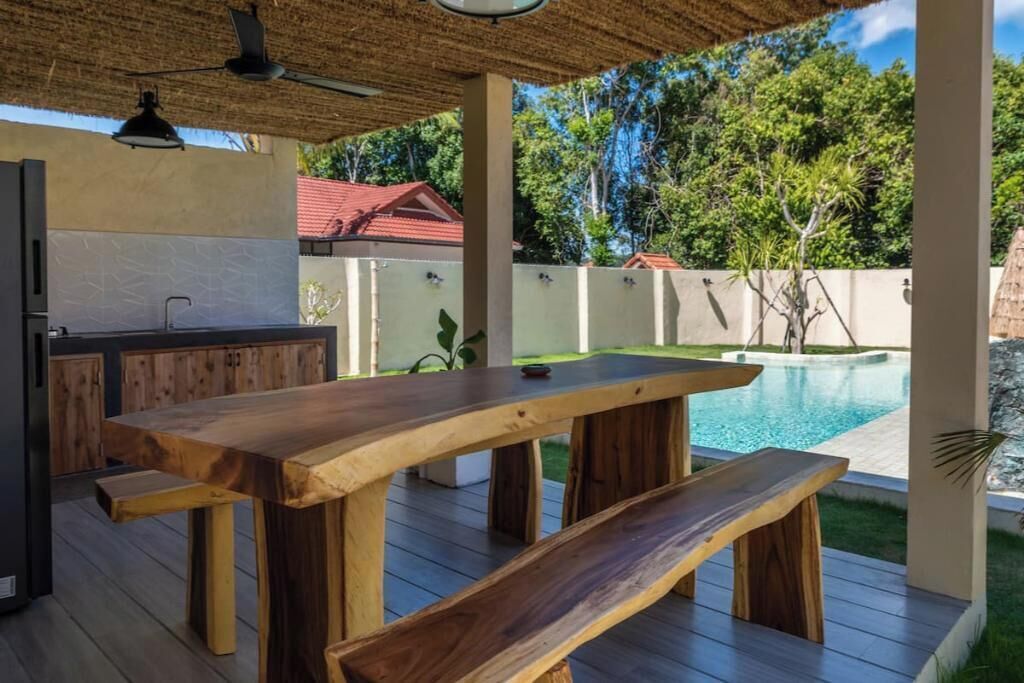 Ein Außenbereich mit einer Holzterrasse, Tischen und Bänken, einem Pool im Hintergrund und einem Reetdach darüber.