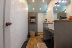 Un baño con suelo de madera, pared de azulejos blancos, lavamanos gris y un espejo sobre el lavamanos.