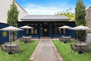 A building with a blue facade, a black door, and a sign with 'OZI' on it. There are four tables with chairs and umbrellas outside. - Private Ensuite Room | Near UniMelb & CBD | Sleeps 3 (Carlton)