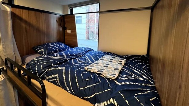 Ein Einzelbett mit einem gemusterten Bettbezug, ein Fenster mit Blick auf Gebäude und ein Holz-Kopfteil.