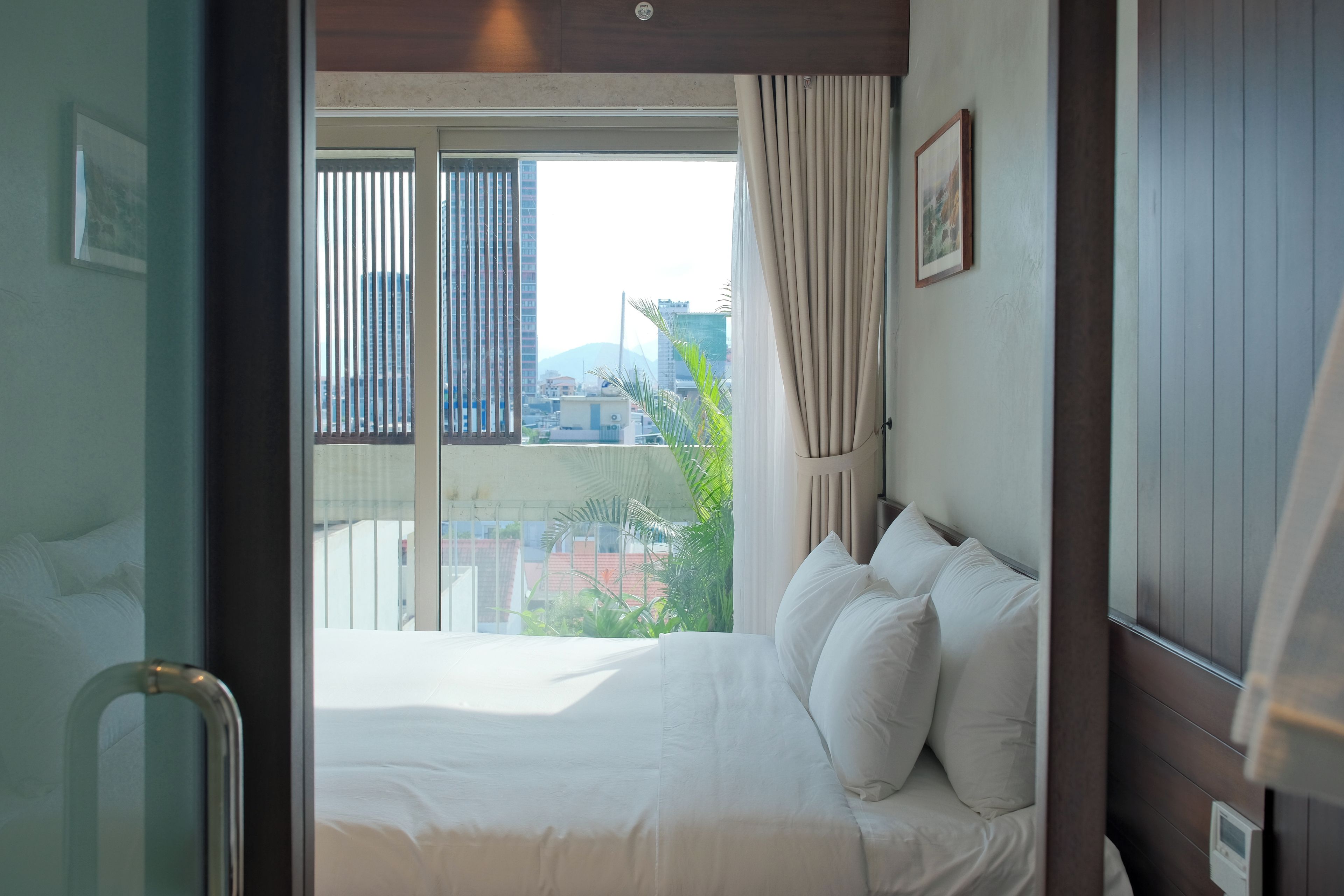 Habitación de hotel con cama, ventana con persianas, cortina y vistas a edificios y áreas verdes.