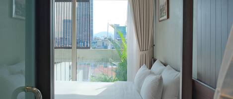 Habitación de hotel con cama, ventana con persianas, cortina y vistas a edificios y áreas verdes.
