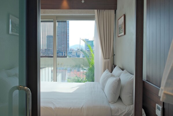 A hotel room with a bed, a window with blinds, a curtain, and a view of buildings and greenery outside. - Marisol Hotel (Da Nang)