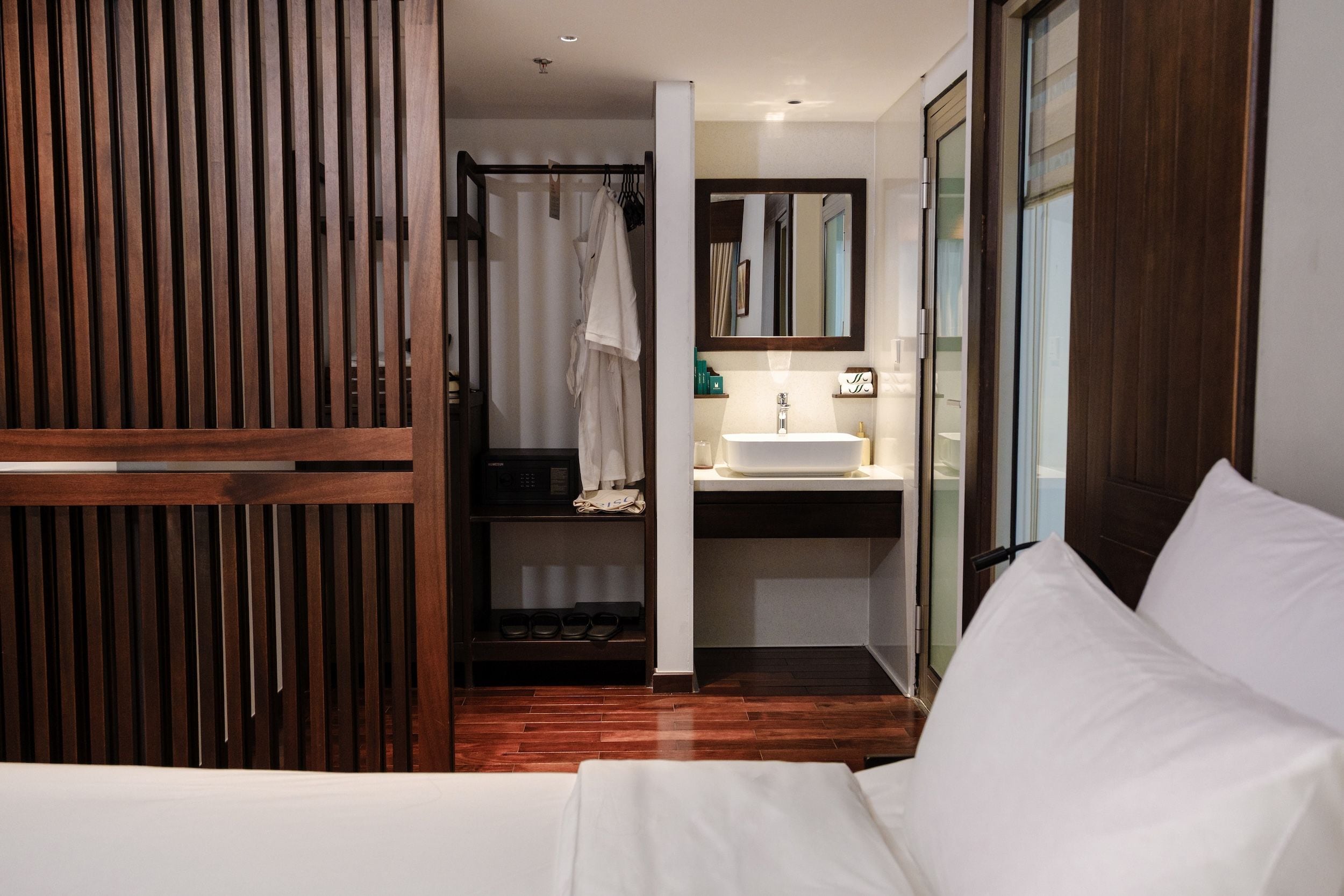 A hotel room with a wooden wardrobe, a bed, a bathroom with a sink and mirror, and a shower area.