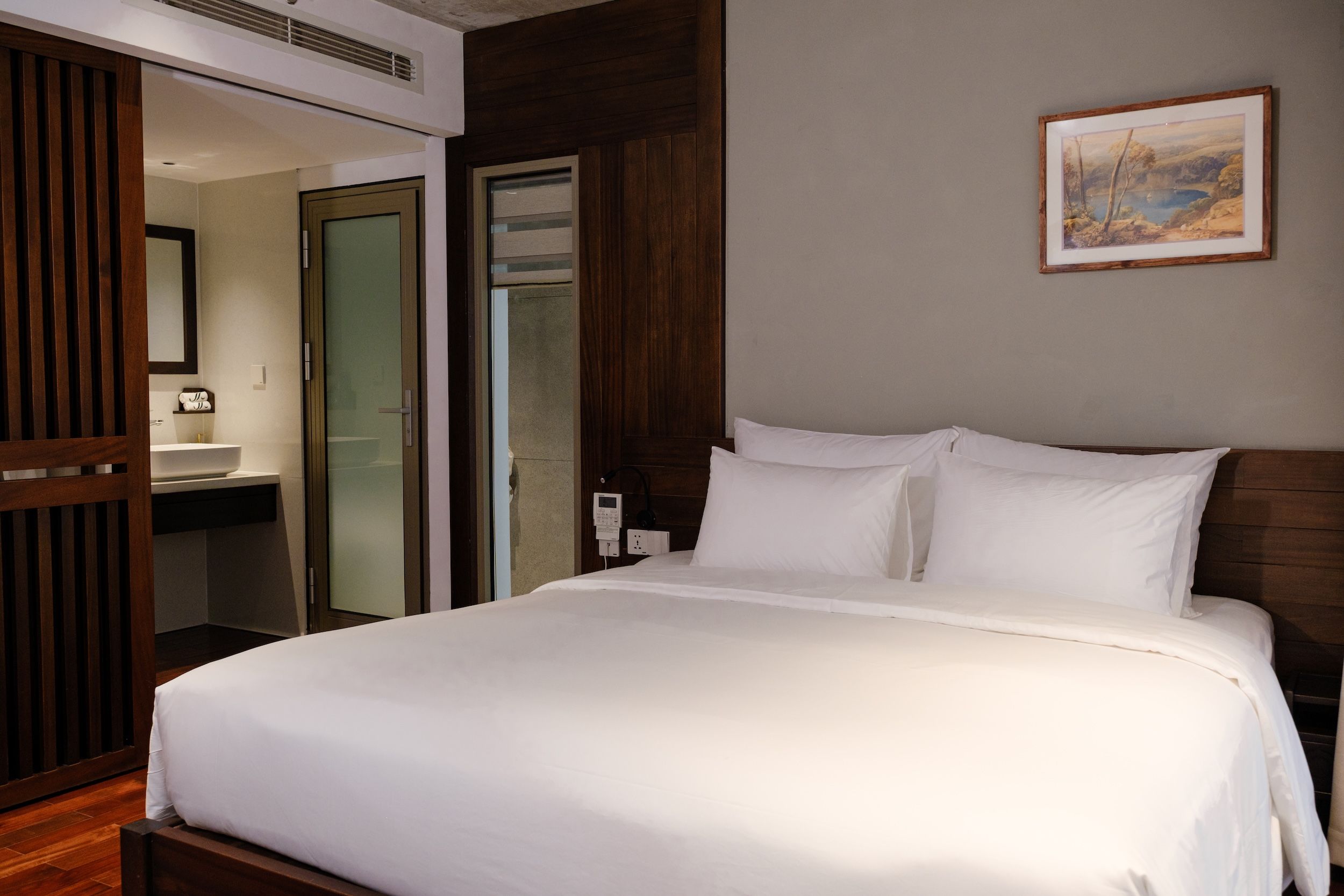 Une chambre d’hôtel avec un grand lit, une tête de lit en bois, un tableau au mur et une salle de bain avec un miroir et un lavabo.